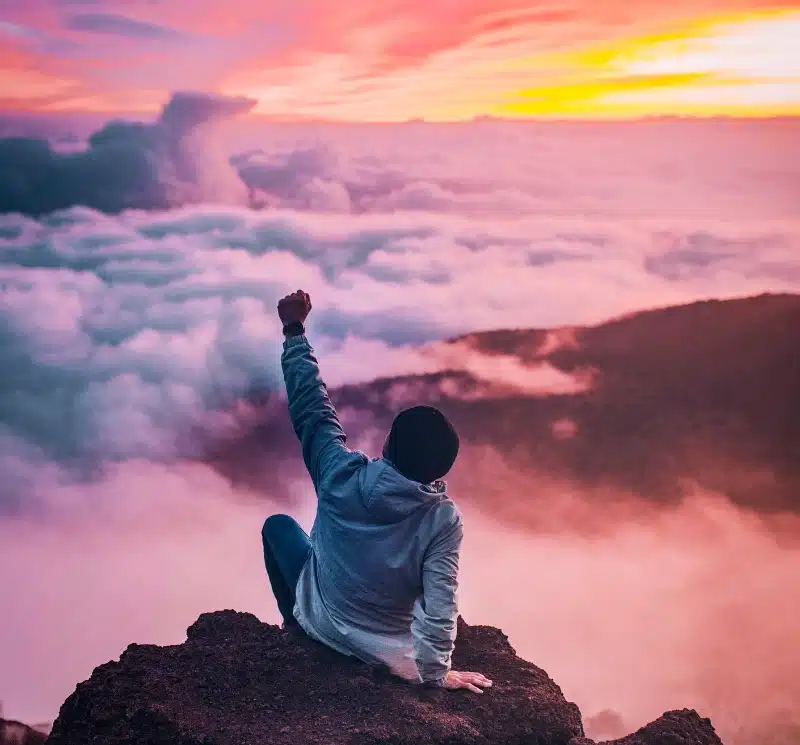 man sitting on mountain cliff facing white clouds rising one hand